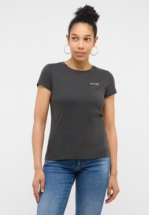 Woman with curly hair in a bun wears gold hoop earrings, a black Mustang t-shirt, and blue jeans, standing against a plain white background.