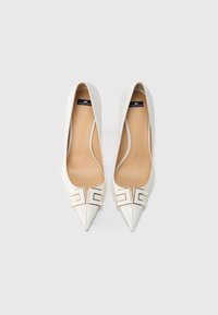 Λευκά δερμάτινα παπούτσια με ψηλό τακούνι και μυτερή μύτη, με χρυσές μεταλλικές λεπτομέρειες και λεία υφή. Η εσωτερική σόλα είναι μπεζ.