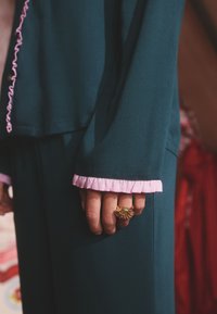 Teal långärmad plagg med rosa volangkantade ärmslut, prydd med en guldring med skaldesign på handen. Mjuk tygstruktur.