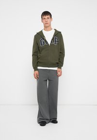 Maslinasto zeleni zip-up dukserica s logotipom "GAP", ispod bijela majica, i sivi široki hlače. Model stoji na jednostavnoj pozadini.