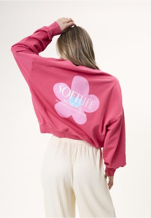 Mujer con cabello rubio que lleva una sudadera rosa con un gran gráfico de flor y texto en la espalda, combinada con pantalones plisados blancos.