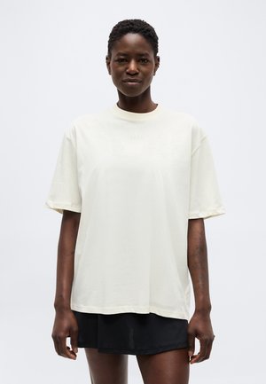Kvinne med kort krøllete hår iført en oversized off-white T-skjorte og svarte shorts, står mot en ensfarget lys bakgrunn.