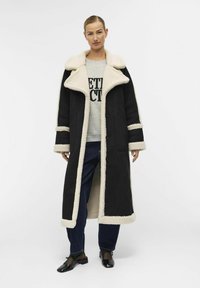 Schwarzer langer Mantel mit einem cremefarbigen Kunstfellkragen und -futter, großen Knöpfen und gerippten Bündchen, kombiniert mit einem grauen Sweatshirt mit Druck und dunklen Jeans.