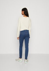 Fehér kötött pulóver, kék farmer skinny cargo nadrág zsebekkel és ezüst sportcipő. Egyszerű dizájn, laza szabású.