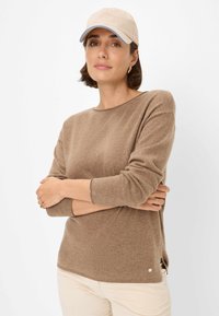 Brauner Pullover mit langen Ärmeln und rundem Ausschnitt, ausgestattet mit einem seitlichen Schlitz und einem kleinen Logo-Detail. Kombiniert mit einer beigen Mütze.