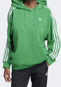 Persoon die een groene Adidas-hoodie draagt met witte strepen op de mouwen en zwarte Adidas-broek met logo op de dij, staand met één hand in de zak.