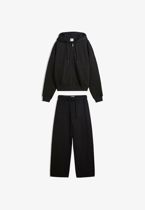 Conjunto casual negro con sudadera con capucha y cremallera, con bolsillos delanteros y pantalones anchos con cordón a juego, extendido plano sobre un fondo blanco.