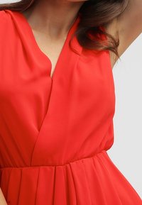 Red sleeveless dress with a v-neckline, featuring soft, smooth fabric, pleated skirt, and a fitted waist. Minimal hardware and detailing.