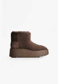 Stivaletti in suede marrone con colletto in pelliccia morbida e suola piatta spessa e testurizzata. Il logo laterale aggiunge un accento notevole.