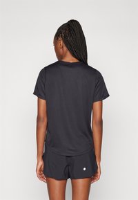 Donna con capelli lunghi intrecciati, indossa una maglietta nera a maniche corte e pantaloncini neri, in piedi con la schiena rivolta verso la fotocamera su uno sfondo neutro.