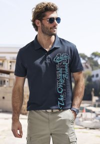 Navy Polohemd mit eine Drei-Knopf-Leiste, versehen mit türkisfarbenem Grafiktext auf der linken Seite. Das Material ist glatt mit einer dezenten Textur.