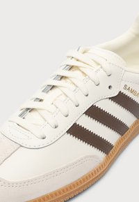 Κοντινό πλάνο λευκού sneaker Adidas Samba με καφέ ρίγες, λευκά κορδόνια, μπεζ καστόρινη μύτη και υφή σόλας σε χρώμα καουτσούκ.