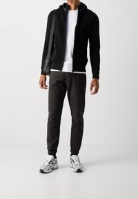 Black zip-up hoodie with a textured fabric, white t-shirt underneath, dark gray tapered pants, and white athletic shoes with silver accents.