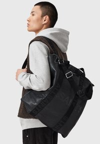 Bolso de cuero negro con superficie texturizada y correa ajustable, con un patrón de logo. El modelo lleva puesta una sudadera gris y un chaleco marrón.
