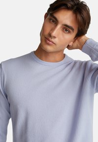 Mladý muž s hnědými vlasy, oblečený v světle fialovém svetru, nakloněný s hlavou a jednou rukou za krkem na jednobarevném pozadí.