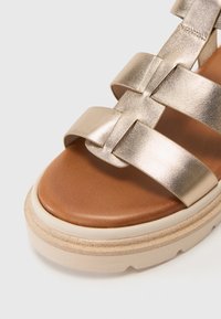 Goldfarbene metallische Riemchensandalette mit einem braunen Lederfußbett und einer chunky cream-farbenen Sohle; verfügt über geflochtene Riemen und eine glatte Textur.