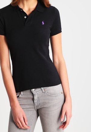 Kvinde iført sort kortærmet polo med lilla logo og lysegrå tætsiddende jeans, stående mod en ensfarvet hvid baggrund.