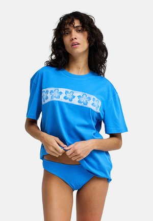 Γυναίκα που φοράει μια φωτεινή μπλε φαρδιά μπλούζα με λευκό floral σχέδιο και ταιριαστά μπλε κάτω μέρος μπικίνι, προσαρμόζοντας τη μπλούζα.