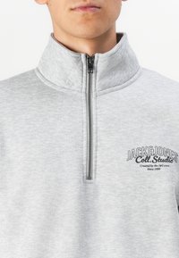 Grå zip-up sweatshirts lavet af blødt stof. Har en høj krave og logoaccent på brystet. Lynlåsen er metal.