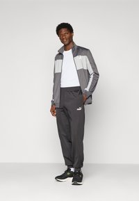 Mand står iført en grå og hvid træningsjakke, hvid skjorte, sorte Puma sweatpants og sorte sportssko med hvide såler.