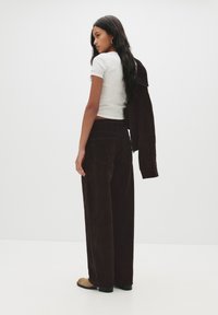 Ruskeat samettihousut, joissa on leveä lahje ja sileä pinta, yhdistettynä istuvaan harmaaseen t-paitaan. Aiheen hiukset ovat pitkät ja laineikkaat.