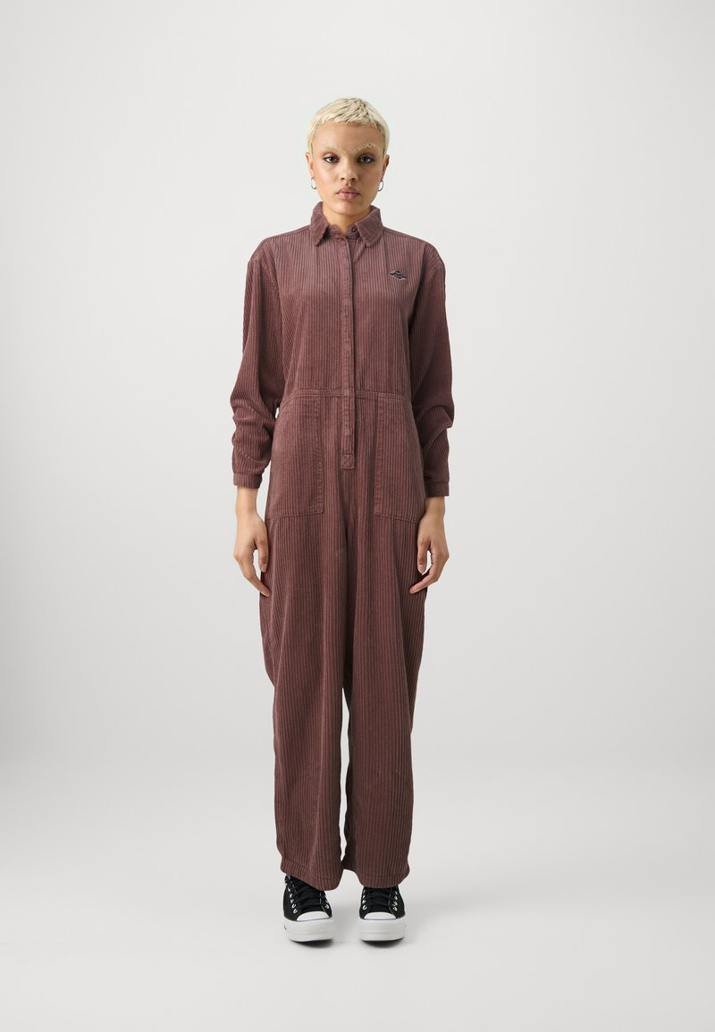 Jumpsuit de pana burdeos con mangas largas, cierre frontal de botones y dos bolsillos delanteros. Combinado con zapatillas negras.
