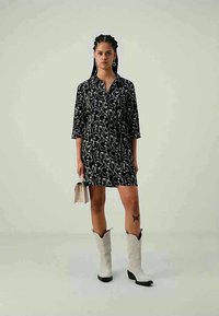 Kobieta z warkoczami, w czarno-białej wzorzystej sukience, w białych kowbojskich butach, trzymająca małą beżową torebkę, stojąca na jasnym tle.