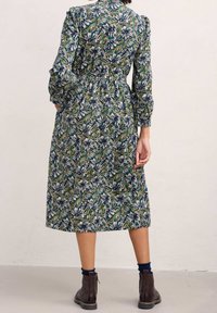 Midi šaty s květinovým vzorem v zelené, modré a bílé; mají dlouhé rukávy, pásek v pase a uvolněný střih. Nosí se s tmavě hnědými kotníkovými botami.