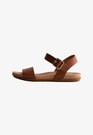 Brown flat sandal with two leather straps, one over toes and one with a gold buckle around the ankle, on a light sole.