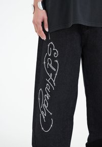 Svarta denimbyxor med rhinestones längs sidan, i lös passform och mjuk textur. Stylandet inkluderar en svart topp.