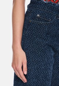 Dunkelblaue, hochtaillierte Hose aus strukturiertem Stoff mit dezentem Muster, vorn mit Tasche und metallischem Akzent am Bund.