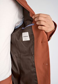 Blazer color óxido con forro marrón liso. La etiqueta interior dice "LINDHBERGH" y "SLIM FIT". Presenta un bolsillo de acento azul en el interior.