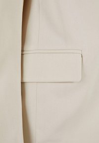 Gros plan sur la poche beige d'une veste sur mesure avec rabat et coutures visibles sur un tissu lisse.