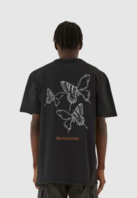 Sort oversized t-shirt med hvide sommerfugle-grafikker og orange tekst "Hvor frygt møder frihed" på ryggen. Blødt bomuldsstof.