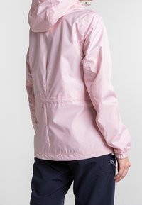 Vaaleanpunainen vedenpitävä takki, jossa on huppu, joustavat hihansuut ja yksinkertaiset sauman yksityiskohdat, yhdistetty tummansinisiin housuihin, esitellen rentoa mallia.