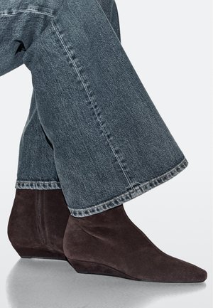 Stivaletti con zeppa in camoscio marrone scuro, punta appuntita e design elegante, abbinati a jeans a gamba larga. Cuciture chiaramente visibili.