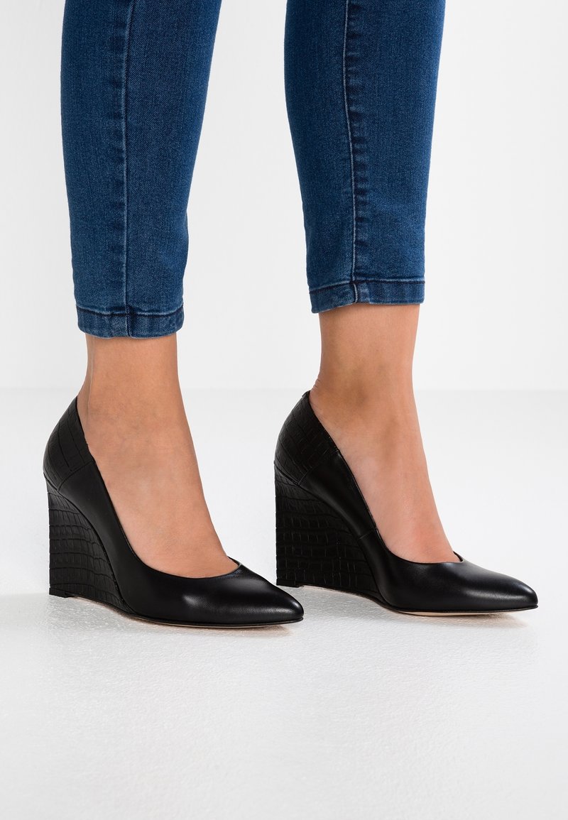 Femme portant des talons compensés en cuir noir à bout pointu avec un motif texturé, associés à un jean skinny bleu foncé raccourci.