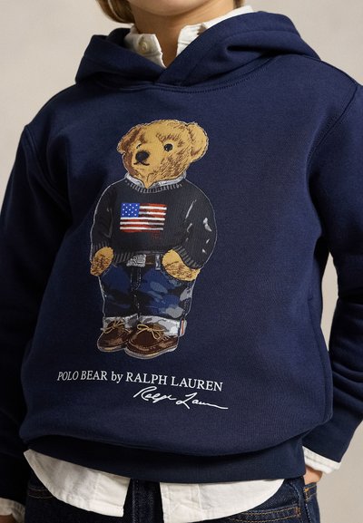 Bambino che indossa una felpa con cappuccio blu navy con grafica di un orso vestito con un maglione con la bandiera americana, pantaloni mimetici e scarpe marroni, etichettata Polo Bear di Ralph Lauren.