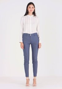 Blå skreddersydde bukser med en glatt tekstur og to frontknapper. Kombinert med en hvit, stripete bluse med lange ermer. Hvite høye sandaler.