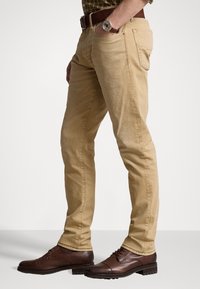 Pantalones chinos de algodón beige con corte ajustado, que cuentan con bolsillos laterales y traseros, combinados con zapatos de cuero marrón oscuro.