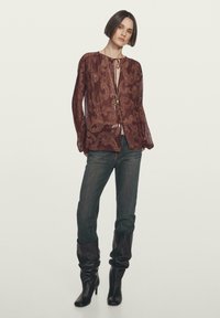 Blusa bordeaux con motivo paisley, maniche lunghe e colletto con fiocco, abbinata a jeans in denim scuro e stivali in pelle nera morbidi.