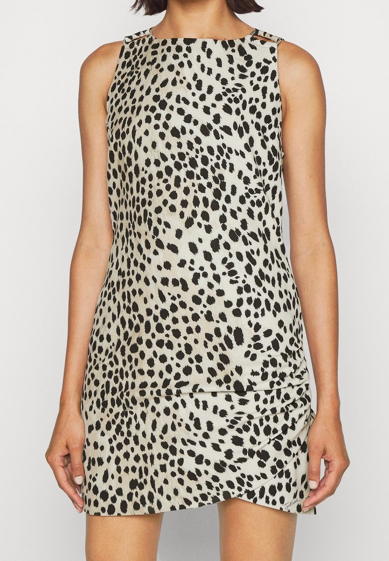 Robe courte sans manches avec des taches noires irrégulières sur un fond beige clair, portée par une femme aux cheveux courts.