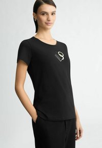 Zwarte t-shirt met korte mouwen en ronde hals, met een klein beige grafisch ontwerp op de borst en een relaxte pasvorm. Soepele textuur.