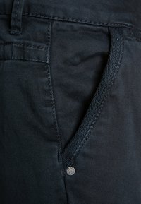 Los pantalones de mezclilla azul oscuro tienen una textura suave, un diseño tradicional de cinco bolsillos y un adorno metálico con el logo en el bolsillo.
