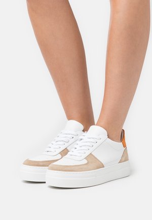 Baskets à plateforme blanches et beiges avec des lacets blancs portées aux pieds sur un fond blanc uni.