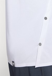 White short-sleeve shirt with a textured finish and silver button accents, featuring a label at the hem. Lightweight, breathable fabric.