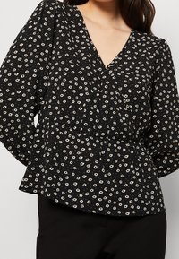 Czarna bluzka z głębokim dekoltem w kształcie litery V, długimi bufkami i dopasowaną talią, ozdobiona białym kwiatowym wzorem i drobnymi groszkami.