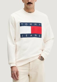 Krämfärgad sweatshirt med en rektangulär grafik som visar texten "TOMMY JEANS", blå och röda accenter samt ribbade muddar och fåll.