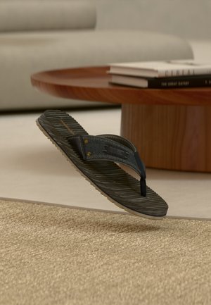Zwarte en grijze teenslipper met een gestructureerde zool, zwevend boven een geweven beige vloerkleed in een moderne woonkamer.