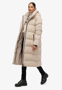 Beige, gewatteerde lange jas met een hoge kraag, rits aan de voorkant, zijzakken en zwarte details. Draag over een gebreide trui met een patroon en donkere broek.